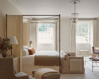Calming, cream bedroom with metal four-poster bed and curtains, seating at end of bed and beside bed, looking towards large windows,